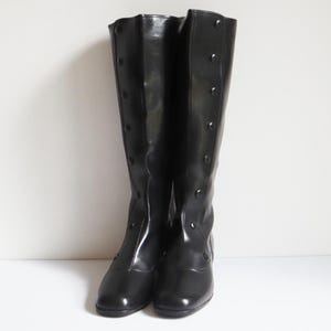 May include: A pair of knee-high black boots featuring a smooth, polished finish and a vertical row of matching black buttons running along the front shaft. The boots have a rounded toe design and a low heel, presenting a sleek and uniform appearance.