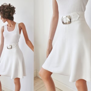 Pode incluir: Saia branca com silhueta evasê e cinto largo a condizer com uma grande fivela prateada. A saia é combinada com um top branco sem mangas. O conjunto é exibido em um manequim.