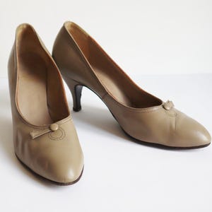 May include: A pair of tan leather court shoes with a small bow detail on the toe. The heels are a classic stiletto style. The shoes are a neutral colour, suitable for various outfits.