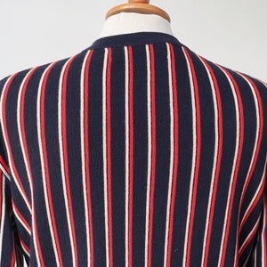 Op de afbeelding: Marineblau gebreide trui met verticale rode en witte strepen. De trui wordt getoond op een mannequin, met de rug en schouder. Het ontwerp is een klassieke, preppy stijl.