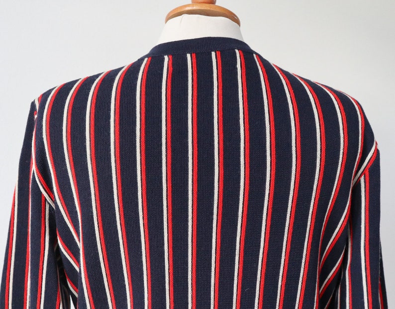 Op de afbeelding: Marineblau gebreide trui met verticale rode en witte strepen. De trui wordt getoond op een mannequin, met de rug en schouder. Het ontwerp is een klassieke, preppy stijl.