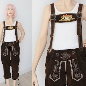 May include: Brown leather lederhosen with suspenders. The lederhosen features decorative embroidery and a crest on the chest. The suspenders are attached with metal buckles. The outfit is worn over a white tank top.