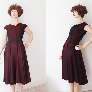May include: A vintage burgundy dress with a fitted bodice and a flared skirt. The dress features short sleeves and a button-up front. The fabric has a subtle sheen, and the dress is displayed on a mannequin. The dress is knee-length.