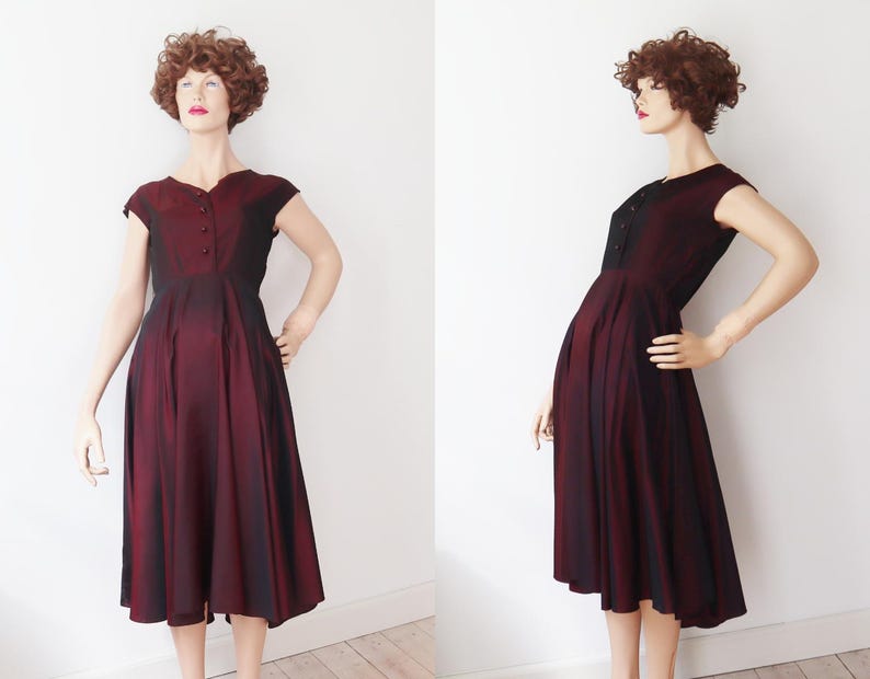 May include: A vintage burgundy dress with a fitted bodice and a flared skirt. The dress features short sleeves and a button-up front. The fabric has a subtle sheen, and the dress is displayed on a mannequin. The dress is knee-length.