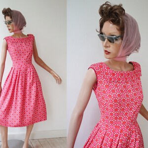 May include: A vintage-style, knee-length dress in a vibrant pink hue, adorned with a repeating geometric pattern in white and red. The dress features a wide, rounded neckline and a pleated skirt, displayed on a mannequin.