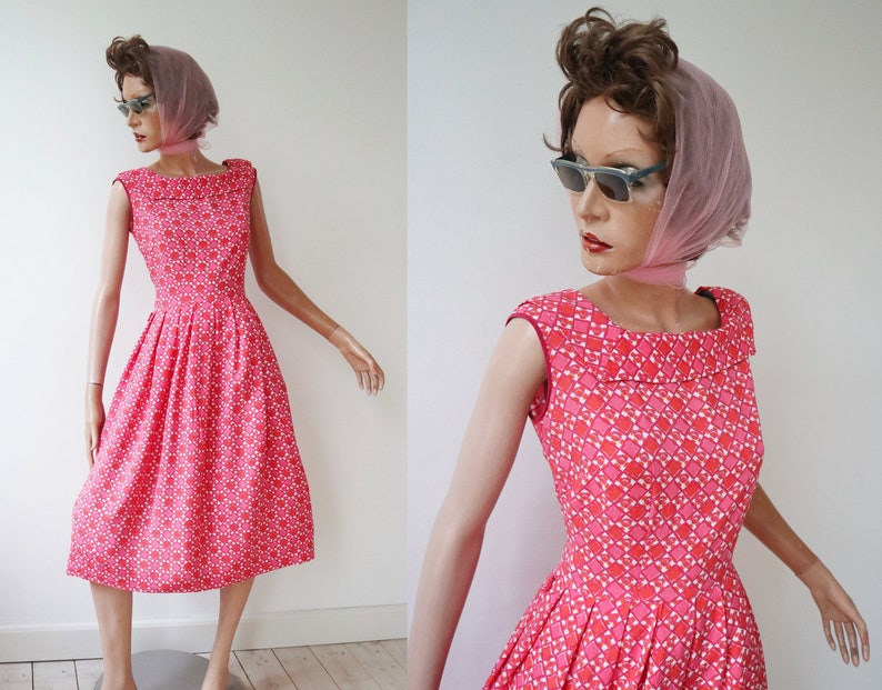 May include: A vintage-style, knee-length dress in a vibrant pink hue, adorned with a repeating geometric pattern in white and red. The dress features a wide, rounded neckline and a pleated skirt, displayed on a mannequin.