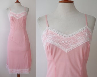 May include: A vintage pink slip dress with delicate white lace trim. The slip features a V-neckline and spaghetti straps. The lace detailing is present on the neckline and hemline, adding a touch of elegance to the garment.