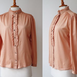 Könnte beinhalten: Pfirsichfarbene Langarmbluse mit hohem Rüschenkragen und vertikaler Rüsche vorne. Die Bluse hat ein dezentes Karomuster und ist vorne geknöpft.