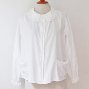 May include: A white long-sleeved blouse with a Peter Pan collar and two front pockets. The collar, cuffs, and pockets are trimmed with delicate lace. The blouse is buttoned down the front and has a classic, feminine design.
