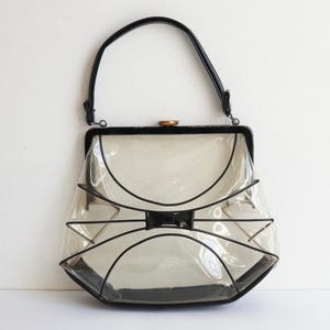Peut inclure: Un sac à main transparent vintage avec des bordures noires et une poignée noire. Le sac présente un design unique en forme de nœud papillon à l'avant et un fermoir doré. Le sac est fait d'un matériau transparent.