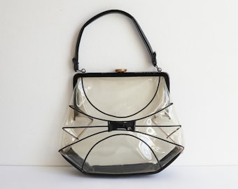 May include: A vintage clear handbag with black trim and a black handle. The bag features a unique bow-shaped design on the front and a gold-colored clasp. The bag is made of a clear, see-through material.