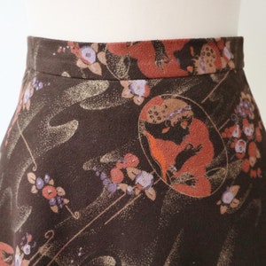K&ouml;nnte beinhalten: A-Linien-Rock mit dunkelbraunem Hintergrund und einem sich wiederholenden Muster aus Blumen- und Figuren-Designs in Orange-, Lila- und Goldt&ouml;nen. Der Rock ist aus einem weichen Stoff gefertigt und hat einen taillierten Bund.