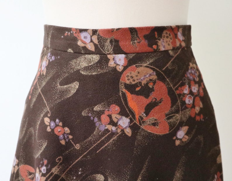 K&ouml;nnte beinhalten: A-Linien-Rock mit dunkelbraunem Hintergrund und einem sich wiederholenden Muster aus Blumen- und Figuren-Designs in Orange-, Lila- und Goldt&ouml;nen. Der Rock ist aus einem weichen Stoff gefertigt und hat einen taillierten Bund.