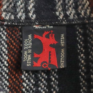 May include: Close-up of a woven fabric with a black, white, and orange plaid pattern. A black label with red text and a dragon emblem reads "WELSH WOOLENS" and "WOVEN IN WALES 100% WOOL".