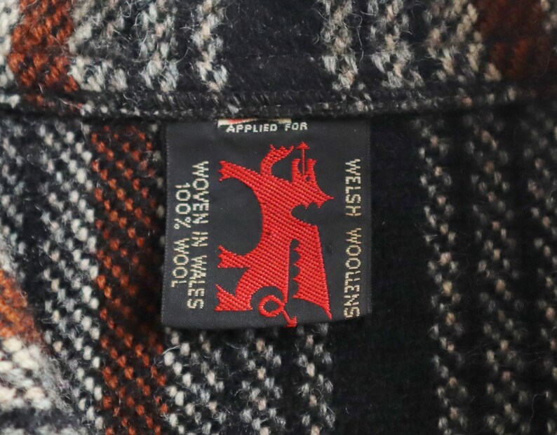 May include: Close-up of a woven fabric with a black, white, and orange plaid pattern. A black label with red text and a dragon emblem reads "WELSH WOOLENS" and "WOVEN IN WALES 100% WOOL".