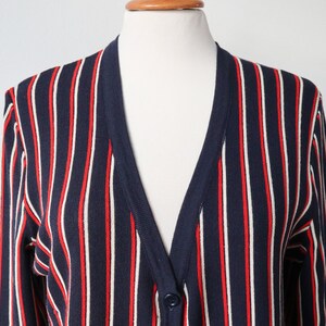 Op de afbeelding: Een marineblauwe cardigan met verticale rode en witte strepen. De V-hals trui heeft lange mouwen en een enkele knoopsluiting. Het kledingstuk wordt op een mannequin gepresenteerd.