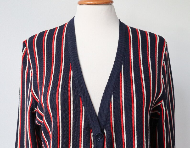 Op de afbeelding: Een marineblauwe cardigan met verticale rode en witte strepen. De V-hals trui heeft lange mouwen en een enkele knoopsluiting. Het kledingstuk wordt op een mannequin gepresenteerd.