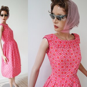 May include: A vintage-style, knee-length dress in a vibrant pink hue, adorned with a repeating geometric pattern in white and darker pink. The dress features a scoop neckline, cap sleeves, and a flared skirt. The dress is displayed on a mannequin.