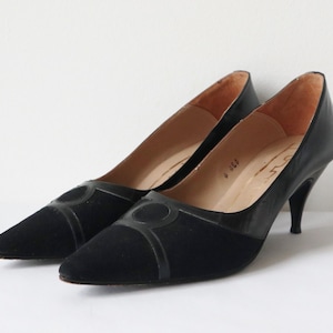 Può includere: Un paio di tacchi neri vintage con punta. Le scarpe presentano un collo del piede in camoscio e dettagli in pelle, con un design circolare decorativo sul davanti. I tacchi sono sottili e curvi.