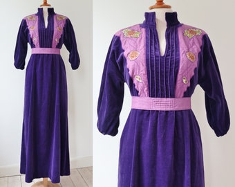 May include: A vintage purple velvet maxi dress with long sleeves and a high collar. The bodice features a light purple panel with floral embroidery and vertical pleats. A matching belt cinches the waist.