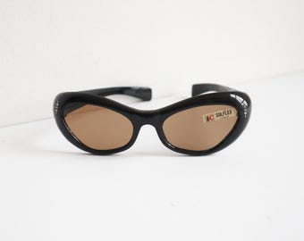 May include: Black cat-eye sunglasses with brown lenses. The sunglasses have a curved frame and small decorative accents near the temples. The label reads "SOLFLEX ITALY".