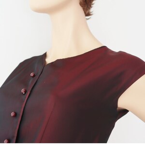 May include: A burgundy-colored, button-down top with short sleeves. The fabric has a subtle sheen, and the buttons match the color of the garment. The top is shown on a mannequin.