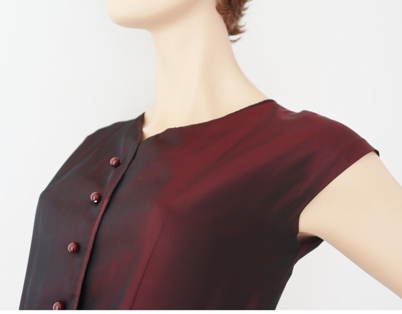 May include: A burgundy-colored, button-down top with short sleeves. The fabric has a subtle sheen, and the buttons match the color of the garment. The top is shown on a mannequin.