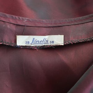 May include: Close-up of a deep burgundy fabric with a label that reads "J&ouml;nells 38". The fabric has a subtle sheen and appears to be part of a garment. The label is rectangular and has black text.