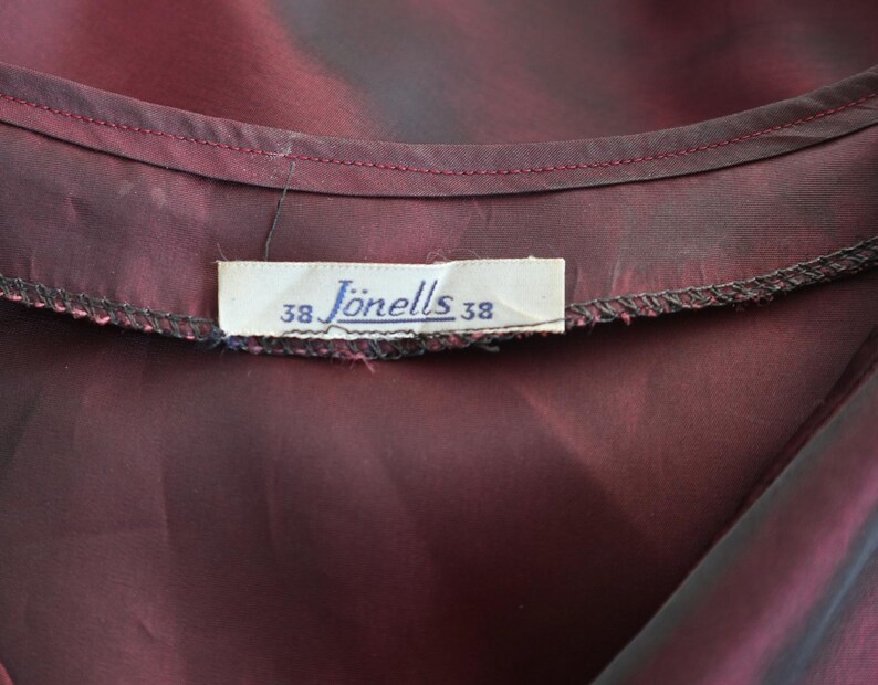 May include: Close-up of a deep burgundy fabric with a label that reads "J&ouml;nells 38". The fabric has a subtle sheen and appears to be part of a garment. The label is rectangular and has black text.