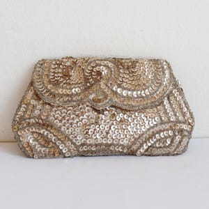 Può includere: Una pochette rettangolare interamente ricoperta di paillettes dorate scintillanti. L'esterno presenta una patta decorativa con bordo smerlato e intricati motivi geometrici formati dalla densa disposizione di piccoli dischi metallici riflettenti.