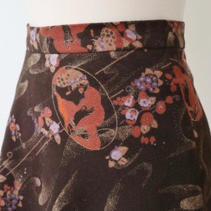 K&ouml;nnte beinhalten: A-Linien-Rock mit dunkelbraunem Hintergrund und einem sich wiederholenden Muster aus floralen und fig&uuml;rlichen Designs in Orange-, Lila- und Beiget&ouml;nen. Der Rock wird auf einer Schaufenquin pr&auml;sentiert.