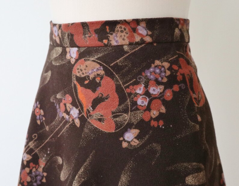 K&ouml;nnte beinhalten: A-Linien-Rock mit dunkelbraunem Hintergrund und einem sich wiederholenden Muster aus floralen und fig&uuml;rlichen Designs in Orange-, Lila- und Beiget&ouml;nen. Der Rock wird auf einer Schaufenquin pr&auml;sentiert.