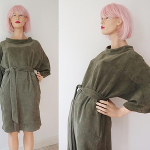 May include: Olive green corduroy dress with a matching belt. The dress features a high neckline, three-quarter sleeves, and a knee-length hemline. The dress is displayed on a mannequin with a pink wig.