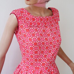 May include: A sleeveless, vintage-style dress featuring a geometric pattern in shades of pink, red, and white. The dress has a square neckline and a flared skirt, showcasing a retro aesthetic. The dress is made of a lightweight fabric.