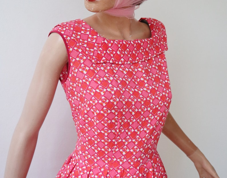May include: A sleeveless, vintage-style dress featuring a geometric pattern in shades of pink, red, and white. The dress has a square neckline and a flared skirt, showcasing a retro aesthetic. The dress is made of a lightweight fabric.