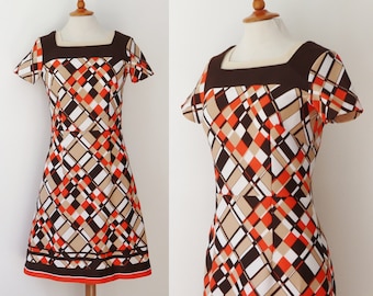 May include: A vintage dress with a geometric pattern. The dress has a square neckline and short sleeves. The pattern includes brown, orange, beige, and white diamond shapes. The hemline is trimmed with a brown and orange border.