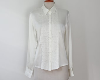 May include: A white long-sleeved shirt with a classic collar and button-down front. The shirt features vertical pleats down the center, adding a textured detail. The cuffs are buttoned, and the shirt is displayed on a mannequin.