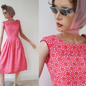 May include: A vintage-style, knee-length dress in a vibrant pink hue, adorned with a repeating geometric pattern of red and white shapes. The dress features a square neckline and a fitted bodice that flares into a full skirt. The mannequin wears sunglasses and a pink scarf.