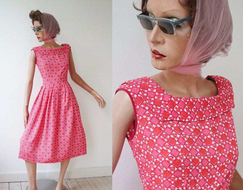 May include: A vintage-style, knee-length dress in a vibrant pink hue, adorned with a repeating geometric pattern of red and white shapes. The dress features a square neckline and a fitted bodice that flares into a full skirt. The mannequin wears sunglasses and a pink scarf.