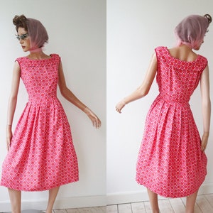 May include: A vintage-style, sleeveless dress in a vibrant red hue, adorned with a repeating white floral pattern. The dress features a fitted bodice, a wide neckline, and a flared skirt, creating a classic silhouette. The dress is displayed on a mannequin.