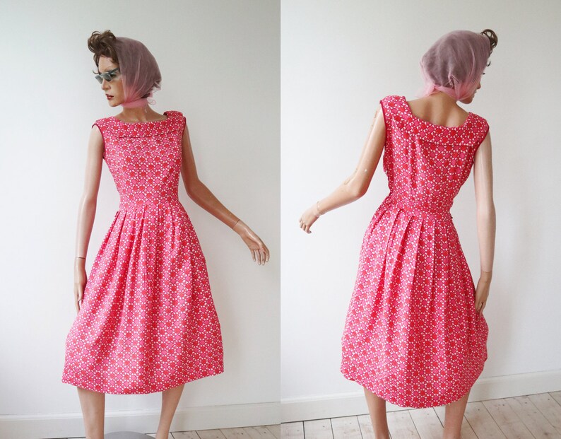 May include: A vintage-style, sleeveless dress in a vibrant red hue, adorned with a repeating white floral pattern. The dress features a fitted bodice, a wide neckline, and a flared skirt, creating a classic silhouette. The dress is displayed on a mannequin.