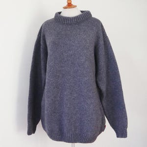 May include: A blue-gray, oversized sweater with a ribbed turtleneck and long sleeves. The sweater is made of a thick, textured knit fabric and is displayed on a mannequin. The garment has a relaxed fit and a classic, timeless design.