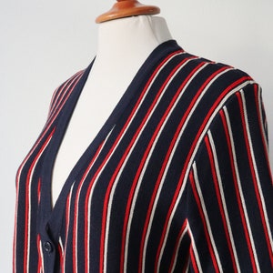 Op de afbeelding: Marineblauwe cardigan met verticale rode en witte strepen. De trui met knopen wordt op een mannequin getoond. De gebreide kleding heeft een V-hals en lange mouwen. Het kledingstuk is gemaakt van een zacht, comfortabel materiaal.