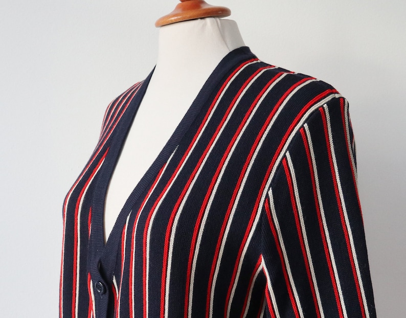 Op de afbeelding: Marineblauwe cardigan met verticale rode en witte strepen. De trui met knopen wordt op een mannequin getoond. De gebreide kleding heeft een V-hals en lange mouwen. Het kledingstuk is gemaakt van een zacht, comfortabel materiaal.