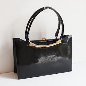 Könnte beinhalten: Eine schwarze Vintage-Handtasche mit glänzendem Finish. Die Tasche hat eine rechteckige Form, einen goldfarbenen Metallbügel oben und zwei schwarze Griffe. Der Verschluss ist ebenfalls goldfarben. Die Tasche ist etwa 25 cm breit.