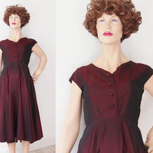 May include: A vintage burgundy dress with a black ombre effect. The dress features a V-neckline, cap sleeves, and a button-down front. The skirt flares out from the waist, creating a classic silhouette. The dress is displayed on a mannequin.