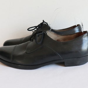 May include: A pair of black leather dress shoes with a classic cap-toe design. The shoes have a low heel and are laced up with black laces. The shoes are displayed on a white background.
