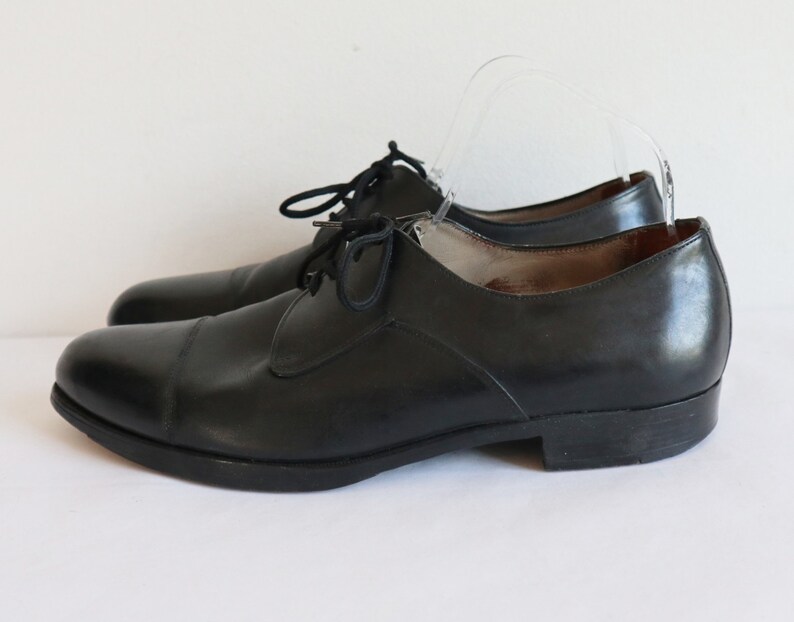 May include: A pair of black leather dress shoes with a classic cap-toe design. The shoes have a low heel and are laced up with black laces. The shoes are displayed on a white background.