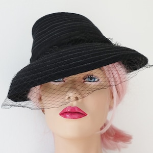 May include: A black hat with a wide brim and a veil. The hat has a structured crown and a textured surface. The veil is a fine black netting that drapes over the front of the hat.
