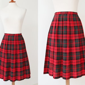 Pode incluir: Saia plissada xadrez vermelha e preta exibida em um manequim. A saia apresenta um padrão de tartan clássico com uma base vermelha e linhas pretas e verdes que se cruzam. A saia é na altura do joelho e tem uma silhueta estruturada em forma de A.